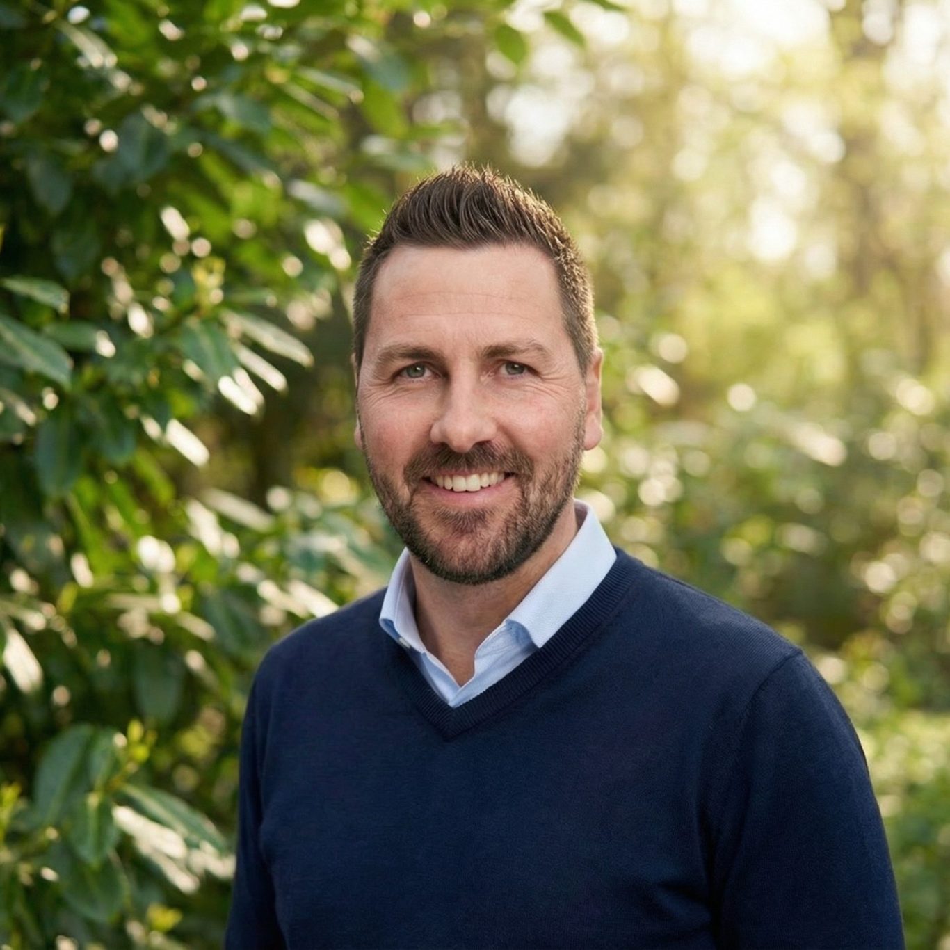 Mann mit Bart in einem blauen Pullover vor einer grünen, sonnigen Hintergrundszene.